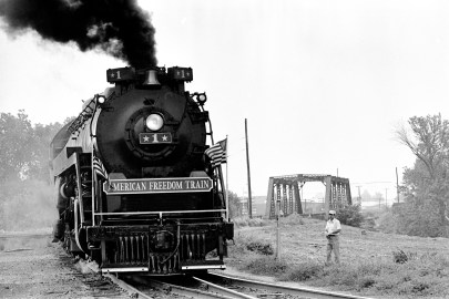 American Freedom Train, steaming around a bend after crossing a bridge. A man watches from the side. "The triumph of the steam-powered American Freedom Train was, indeed, the only nationwide celebration of the Bicentennial. It was pulled by steam locomotives in the age of the diesel, and would improve on the three display cars of its predecessor, the 1947 Freedom Train. The American Freedom Train would feature twelve display cars, ten that visitors would go aboard and pass through and two to hold large objects that would be viewed from the ground through huge "showcase" windows. The display cars were filled with over 500 precious treasures of Americana. Included in these diverse artifacts were George Washington's copy of the Constitution, the original Louisiana Purchase, Judy Garland's dress from The Wizard of OZ, Joe Frazier's boxing trunks, Martin Luther King's pulpit and robes, and even a rock from the moon. The American Freedom Train (AFT) was a 26-car train led by one of three enormous steam engines restored just for the occasion. Over a 21 month period from April 1, 1975 to December 31, 1976 more than 7 million Americans visited the train during its tour of all 48 contiguous states. Tens of millions more stood trackside to see it go by." (http://www.freedomtrain.org/american-freedom-train-home.htm) The Freedom Train's only visit to Evansville was June 15-17, when it was parked behind the Civic Center/Vanderburgh Auditorium at 715 Locust St., 1976. Source: Gregory Smith Collection (MSS 034-0594)