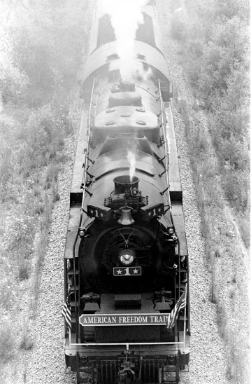 Looking down on the American Freedom Train, steaming down the tracks. "The triumph of the steam-powered American Freedom Train was, indeed, the only nationwide celebration of the Bicentennial. It was pulled by steam locomotives in the age of the diesel, and would improve on the three display cars of its predecessor, the 1947 Freedom Train. The American Freedom Train would feature twelve display cars, ten that visitors would go aboard and pass through and two to hold large objects that would be viewed from the ground through huge "showcase" windows. The display cars were filled with over 500 precious treasures of Americana. Included in these diverse artifacts were George Washington's copy of the Constitution, the original Louisiana Purchase, Judy Garland's dress from The Wizard of OZ, Joe Frazier's boxing trunks, Martin Luther King's pulpit and robes, and even a rock from the moon. The American Freedom Train (AFT) was a 26-car train led by one of three enormous steam engines restored just for the occasion. Over a 21 month period from April 1, 1975 to December 31, 1976 more than 7 million Americans visited the train during its tour of all 48 contiguous states. Tens of millions more stood trackside to see it go by." (http://www.freedomtrain.org/american-freedom-train-home.htm) The Freedom Train's only visit to Evansville was June 15-17, when it was parked behind the Civic Center/Vanderburgh Auditorium at 715 Locust St. in Evansville, Indiana, 1976. Source: Gregory Smith Collection, MSS 034-0604.